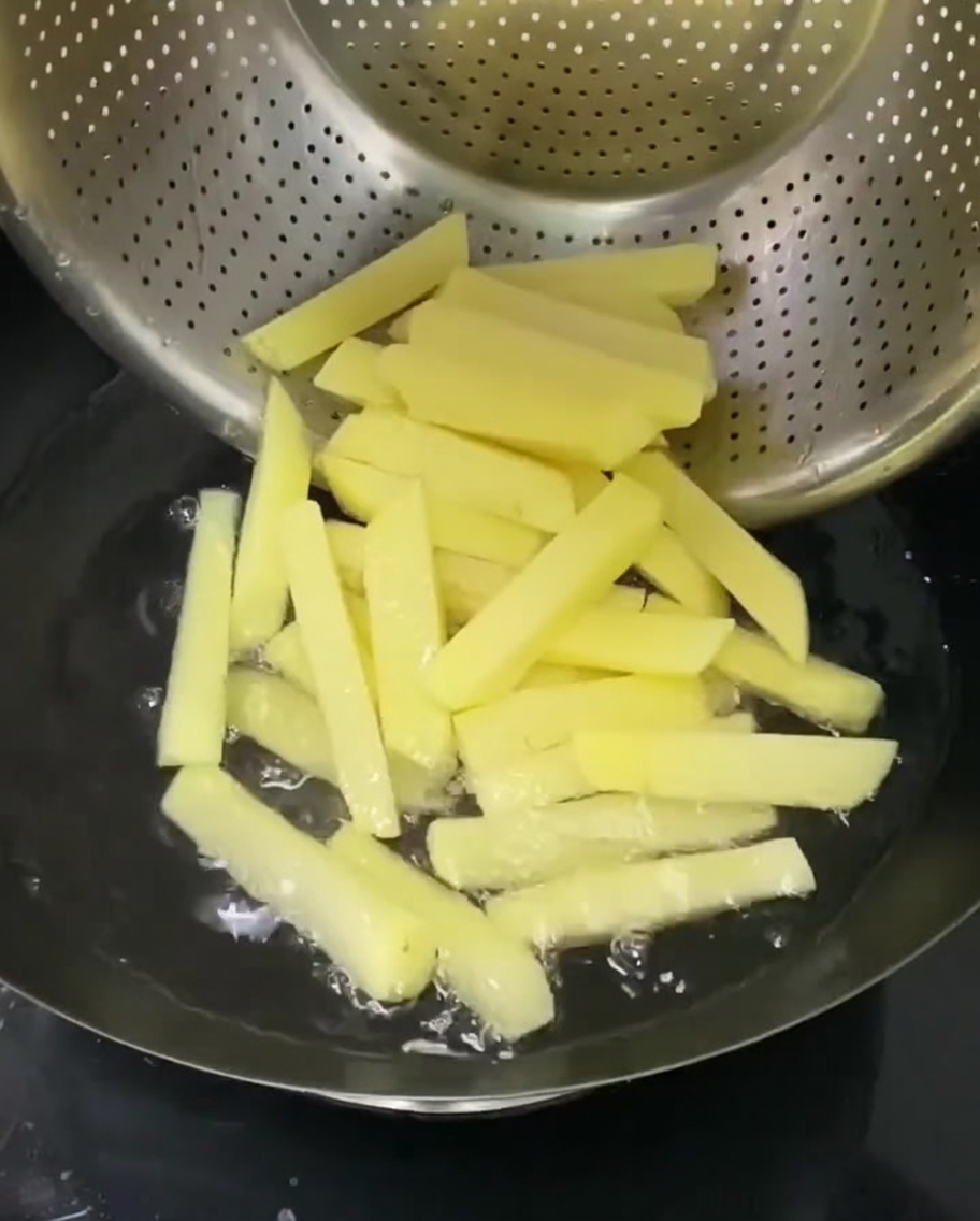 Potato sticks being boiled in a metal colander submerged in a pot of hot water.