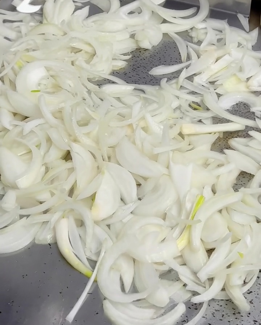 Melted butter being poured from a small saucepan over a pile of onions on a hot griddle.