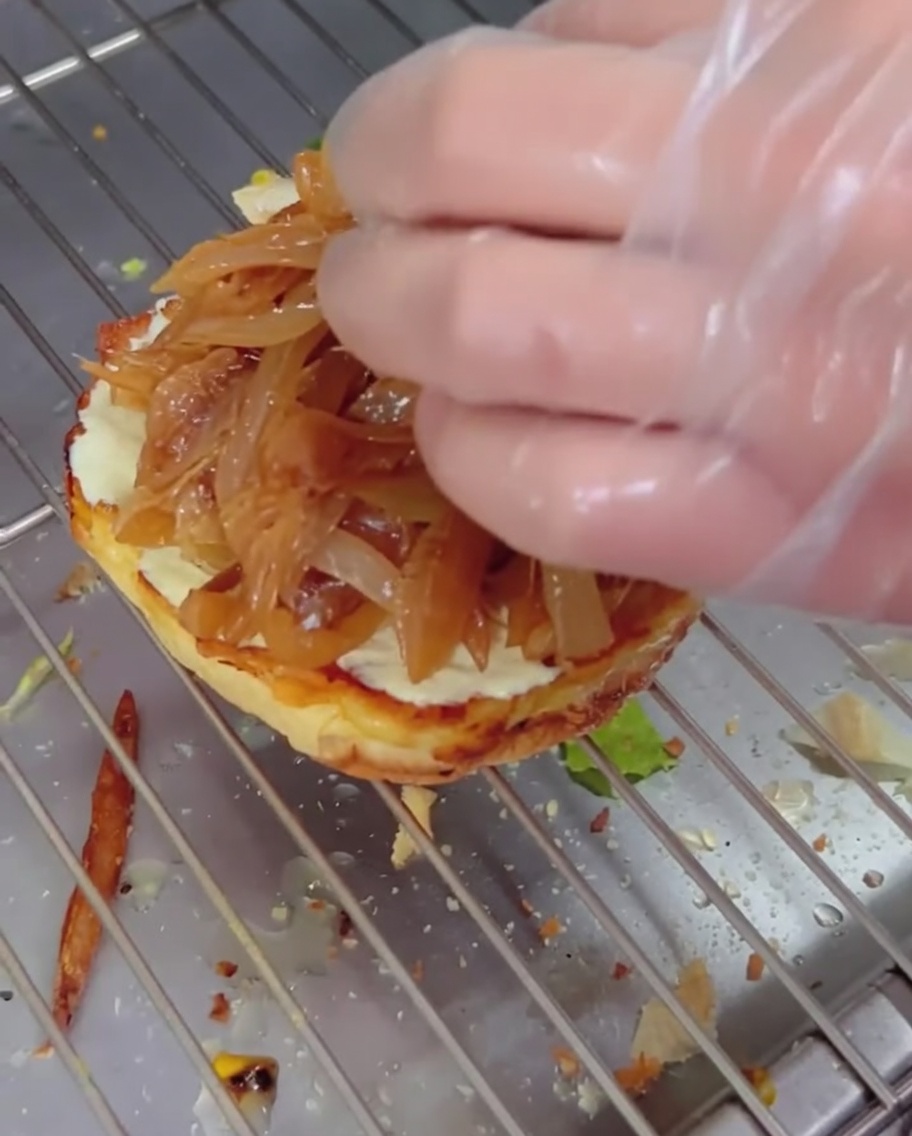 Caramelized onions being layered onto a burger bun with a cheesy beef patty stacked on top.