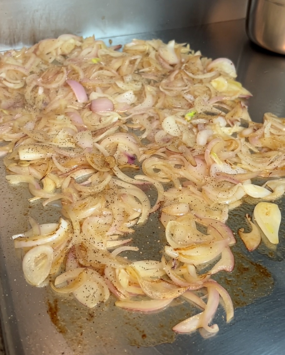 Caramelizing onions on a griddle being seasoned with a sprinkle of black pepper and vibrant Hungarian sweet paprika.