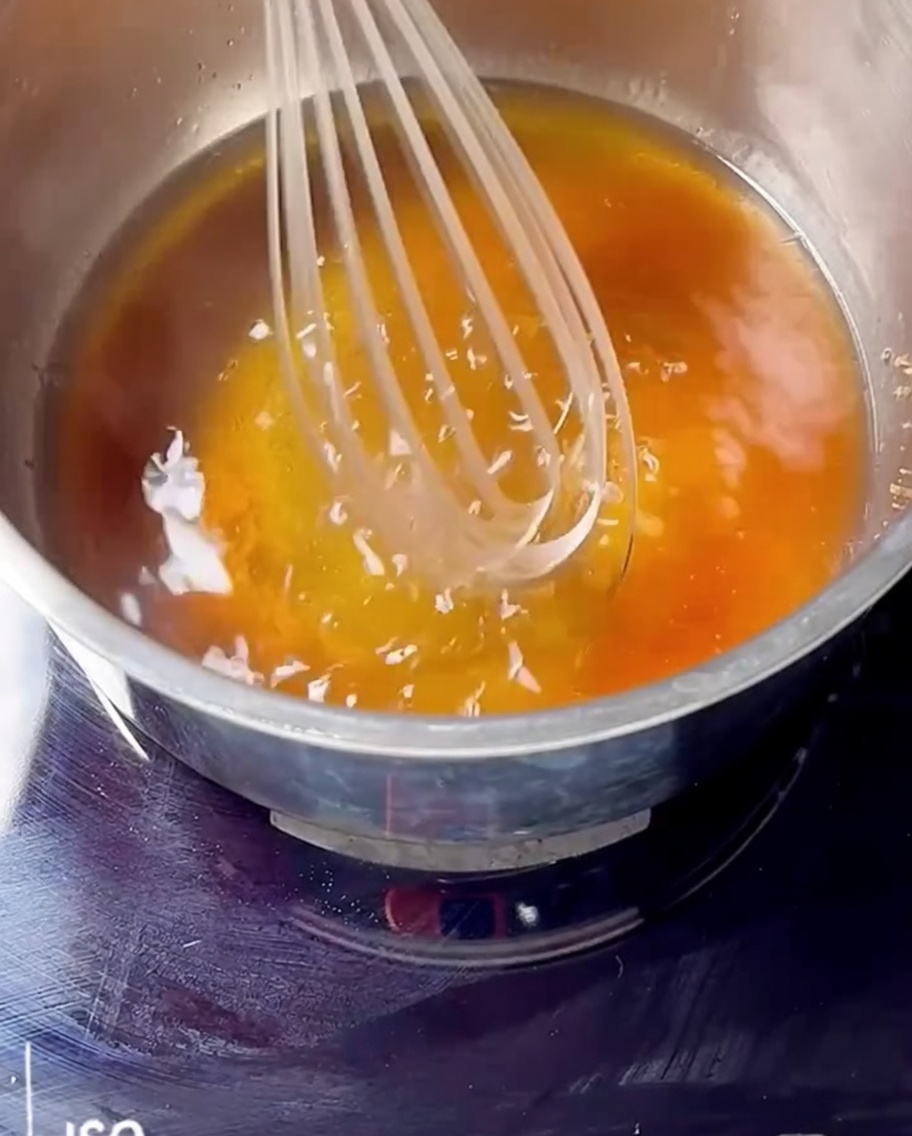A whisk mixing cherry jam, bourbon, and brown sugar in a saucepan on a stovetop.