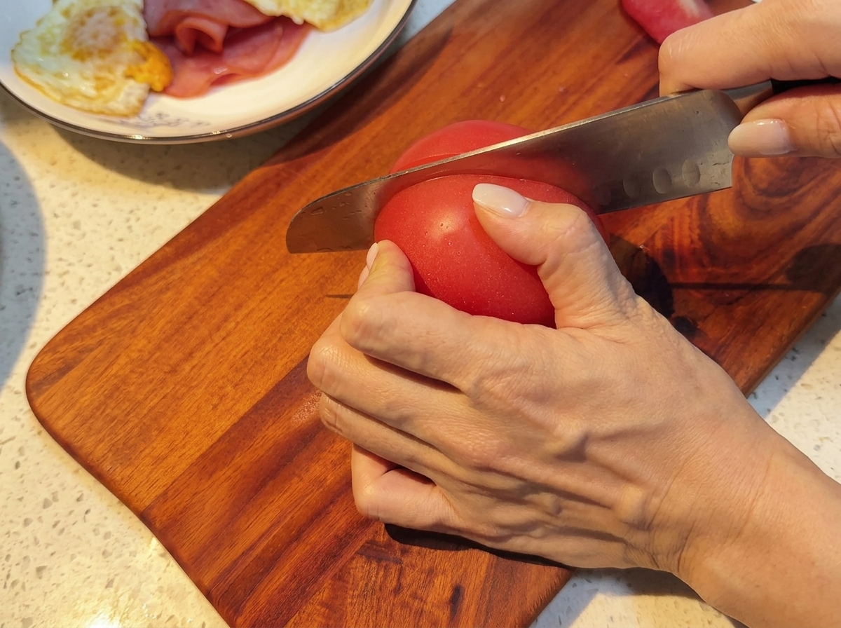 Fresh tomatoes being sliced into uniform rounds on a wooden board.