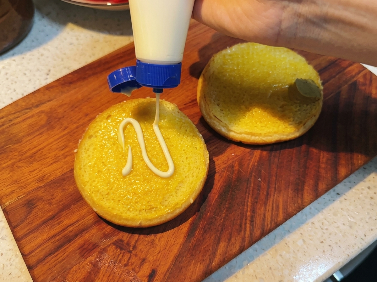 Mayonnaise being squeezed onto a bun already layered with honey mustard.