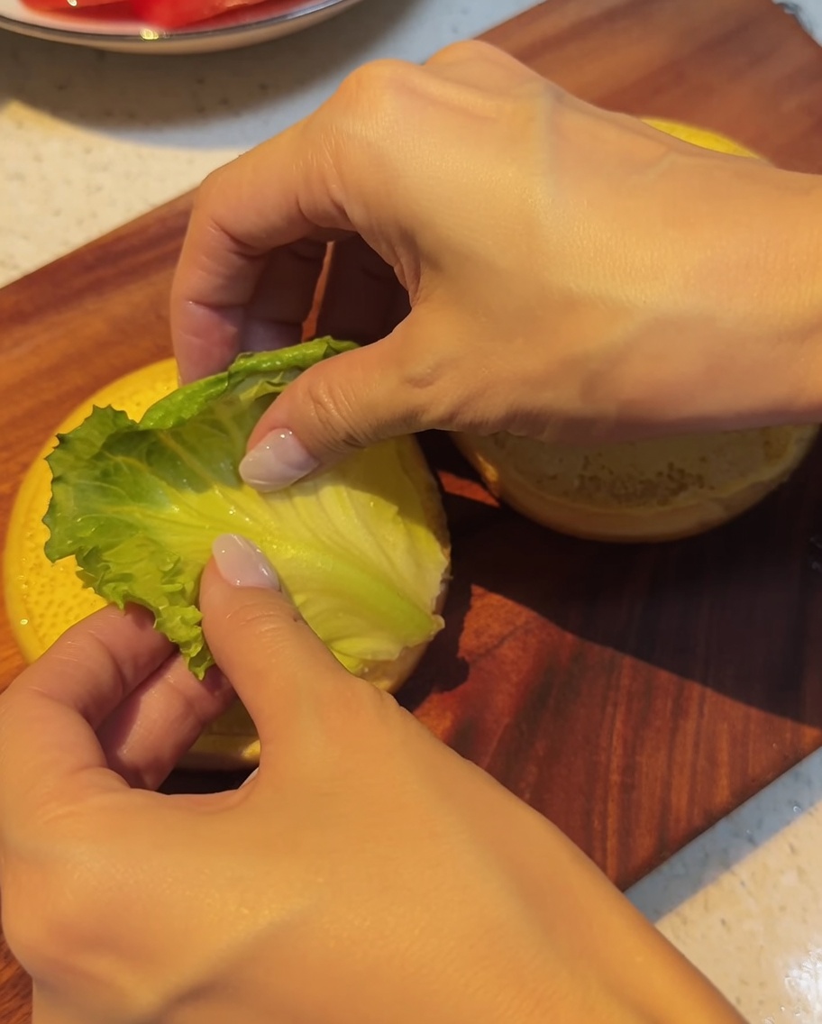 Fresh green lettuce leaves being carefully layered onto the bottom bun on a wooden cutting board.