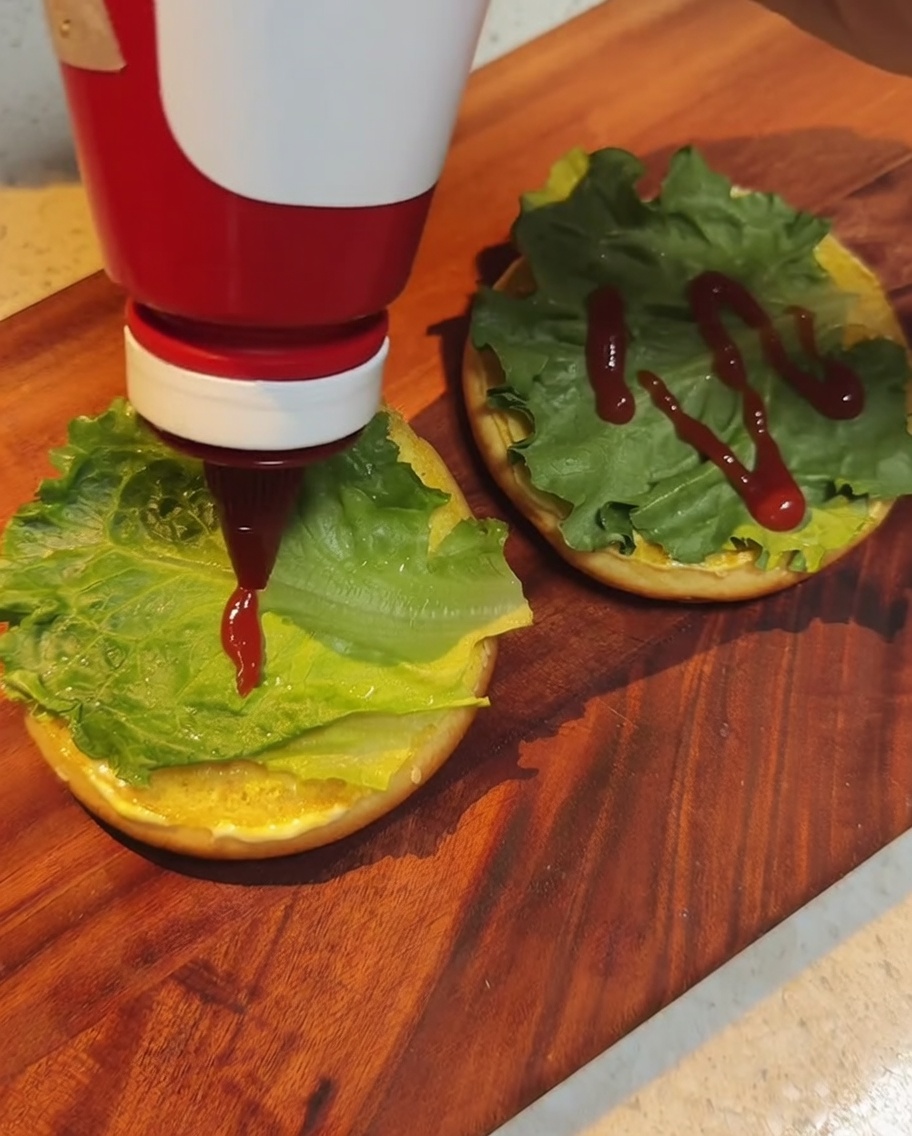 Red ketchup being drizzled from a squeeze bottle over green lettuce leaves on the hamburger bun.