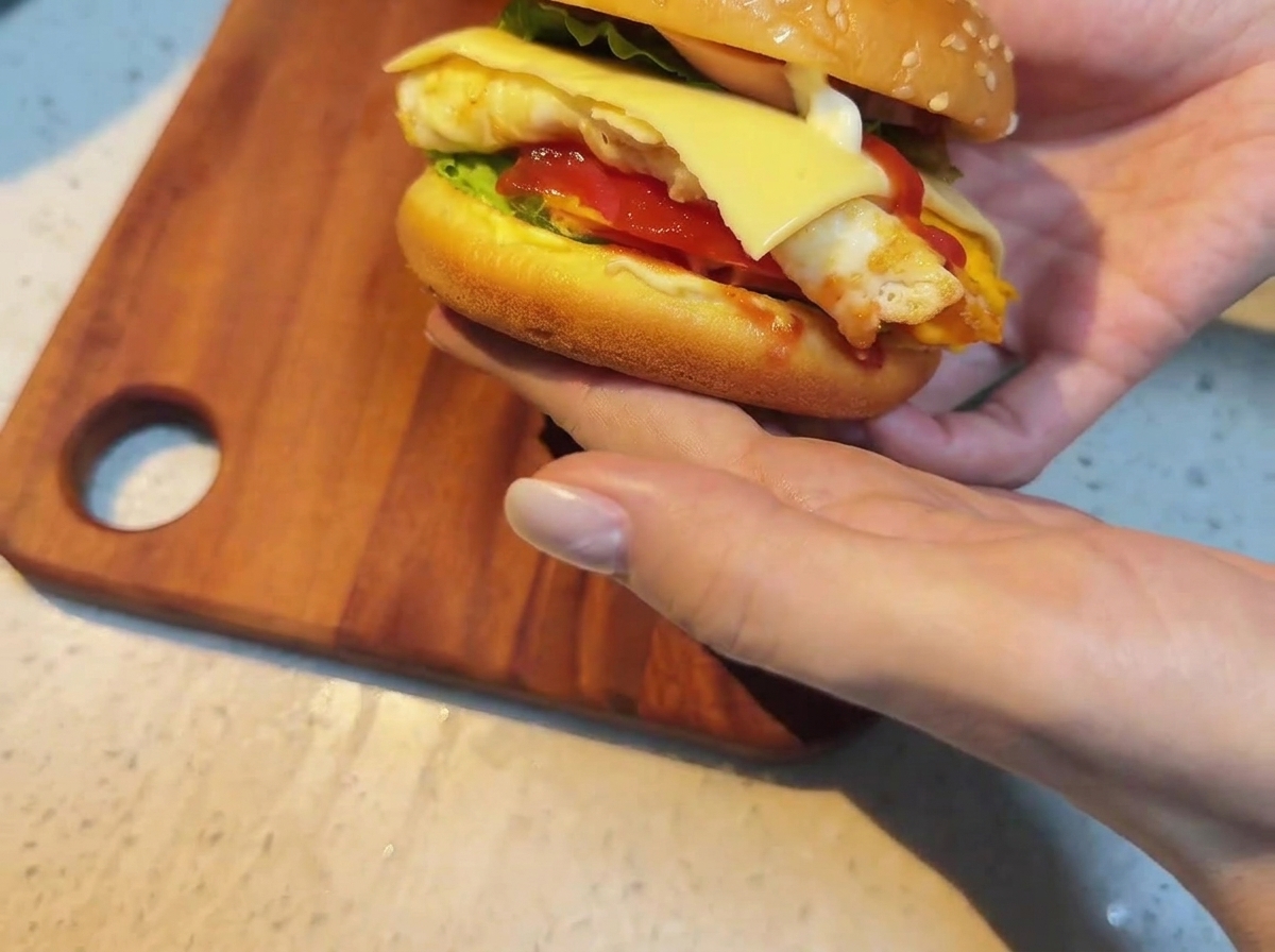 A single slice of cheese being placed on top of the stacked burger ingredients on a wooden board.