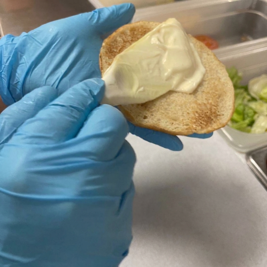 A plastic spreader applying a layer of creamy mayonnaise onto the toasted surface of a burger bun.