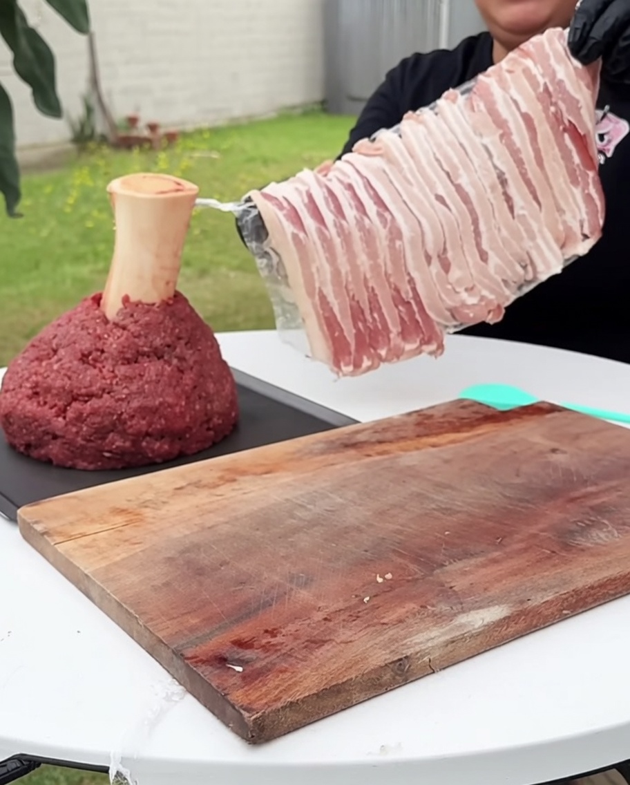 A square block of ground beef being carefully wrapped with strips of raw bacon on all sides.