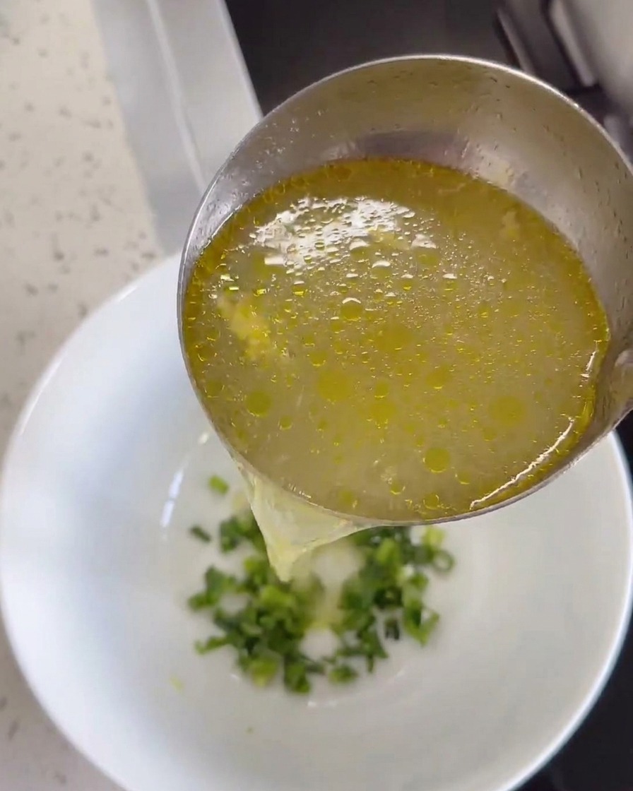 Hot chicken broth being poured into a bowl containing ginger and cilantro.