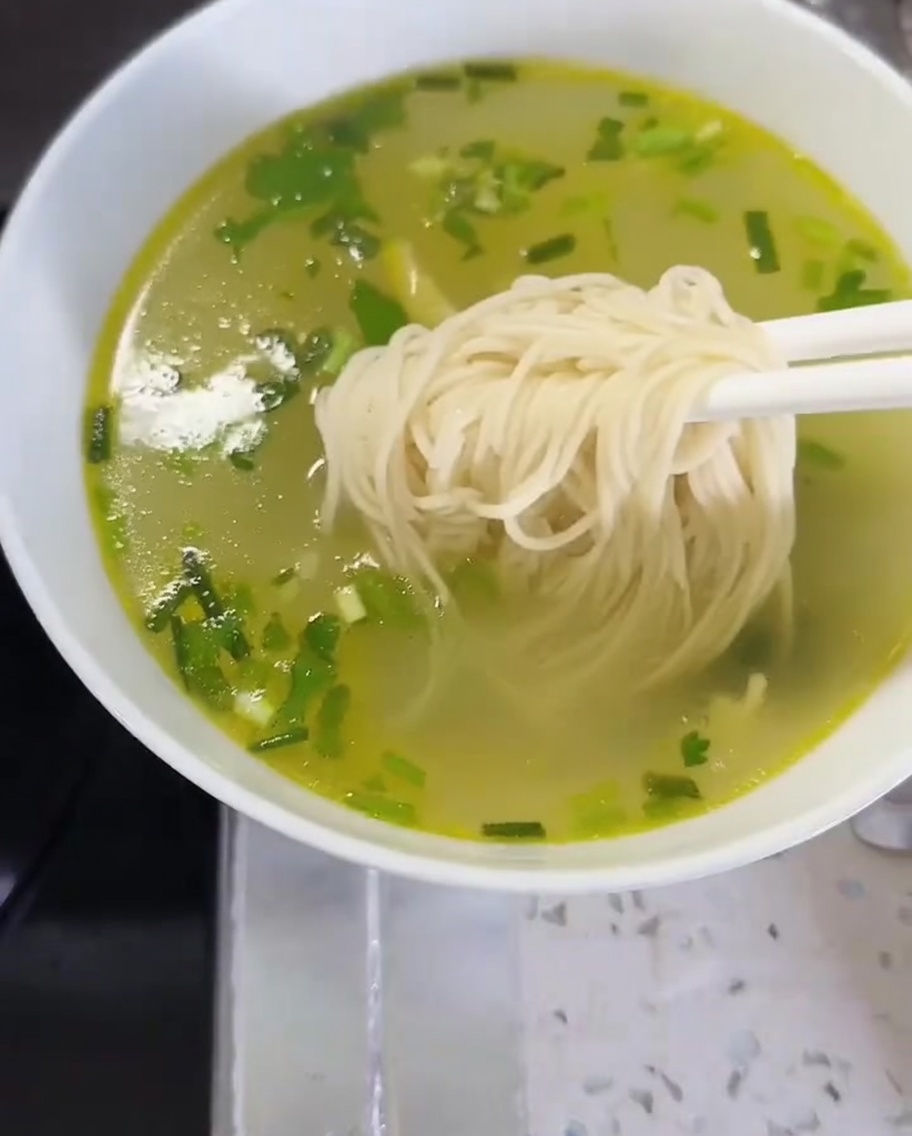 Noodles and greens being transferred into a bowl of broth to complete the dish.