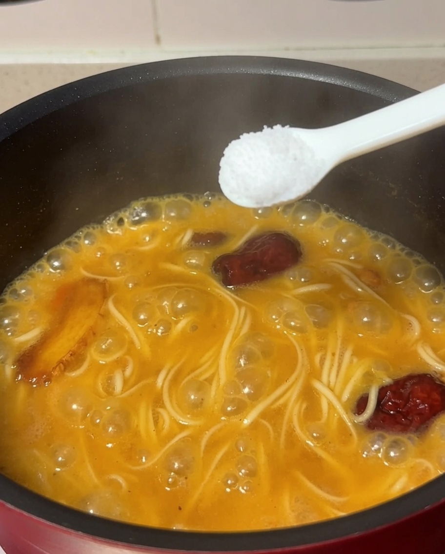 A spoonful of salt being added to the pot containing boiling noodles and chicken soup.