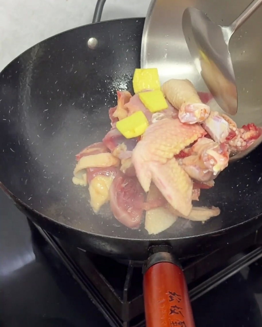 Raw chicken pieces and ginger slices being dropped into a hot black wok for stir-frying.