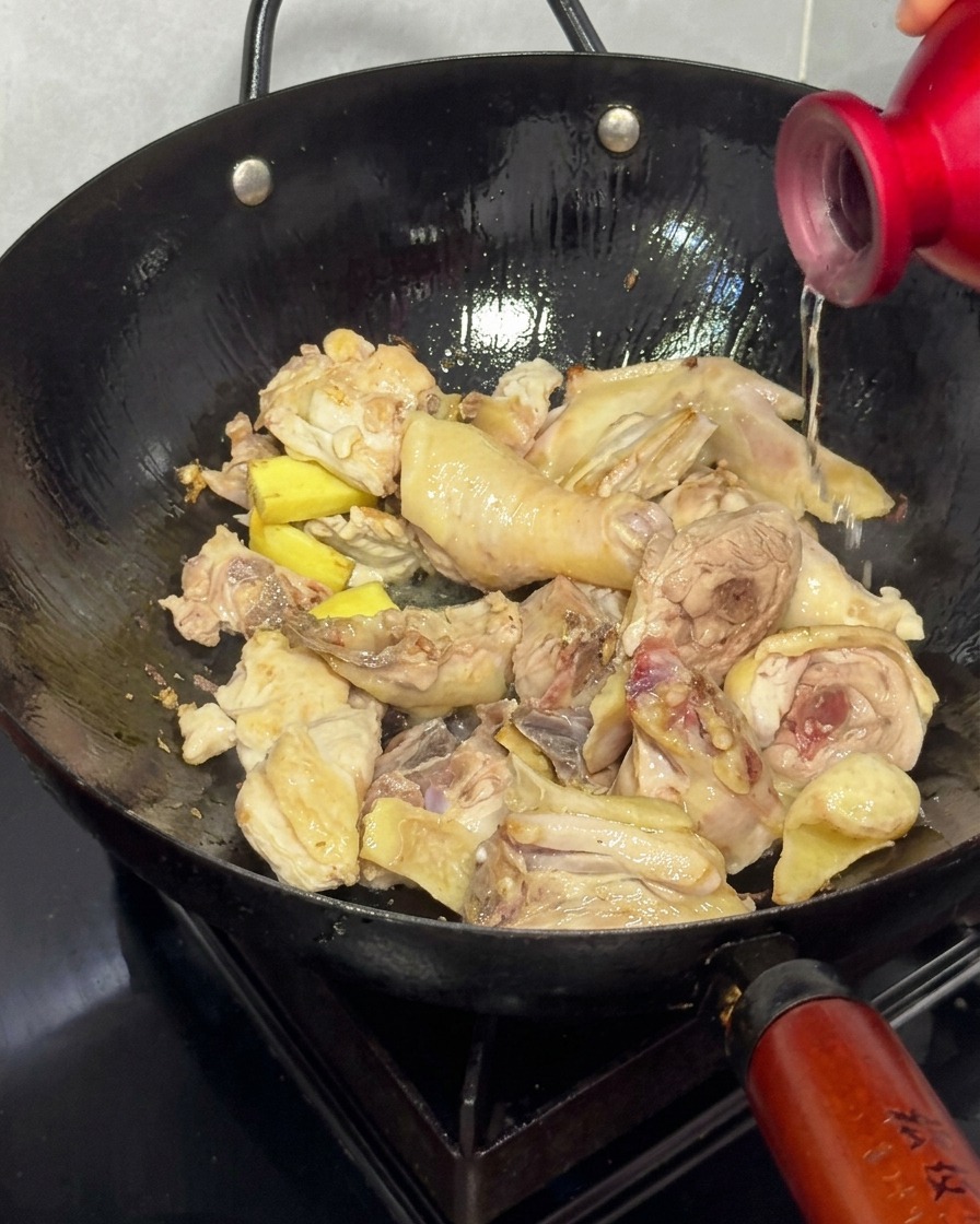 Pouring rice wine from a red bottle into a wok filled with browned chicken pieces and ginger.