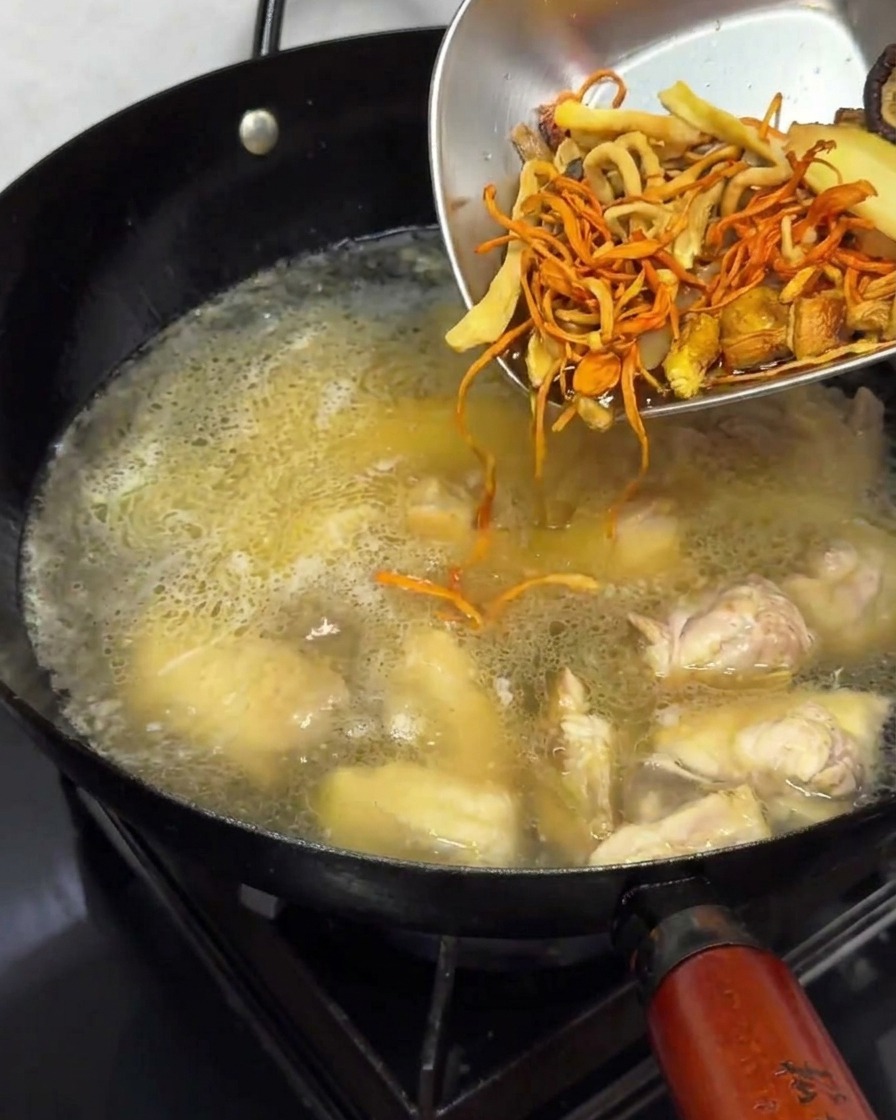 Pouring soaked dried mushrooms from a metal dish into the simmering chicken broth.