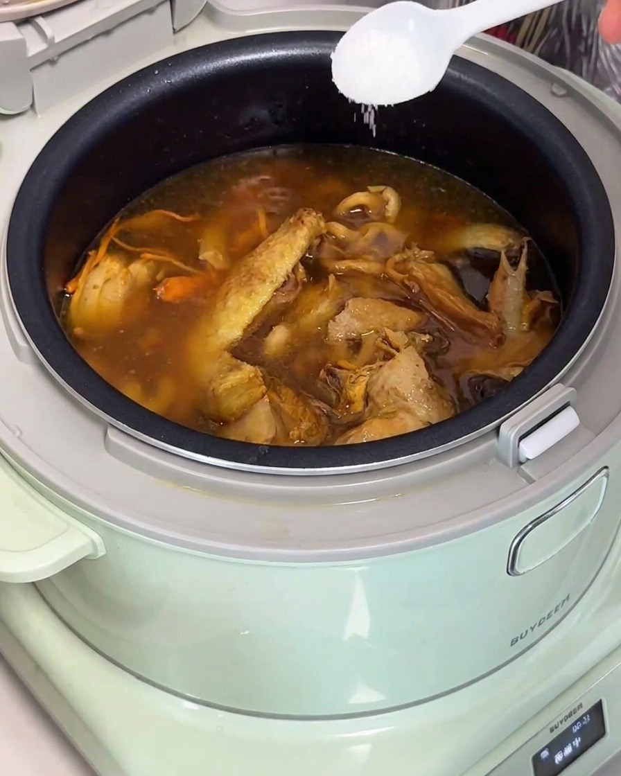A white spoon adding salt into a finished pot of golden chicken and mushroom soup in a rice cooker.