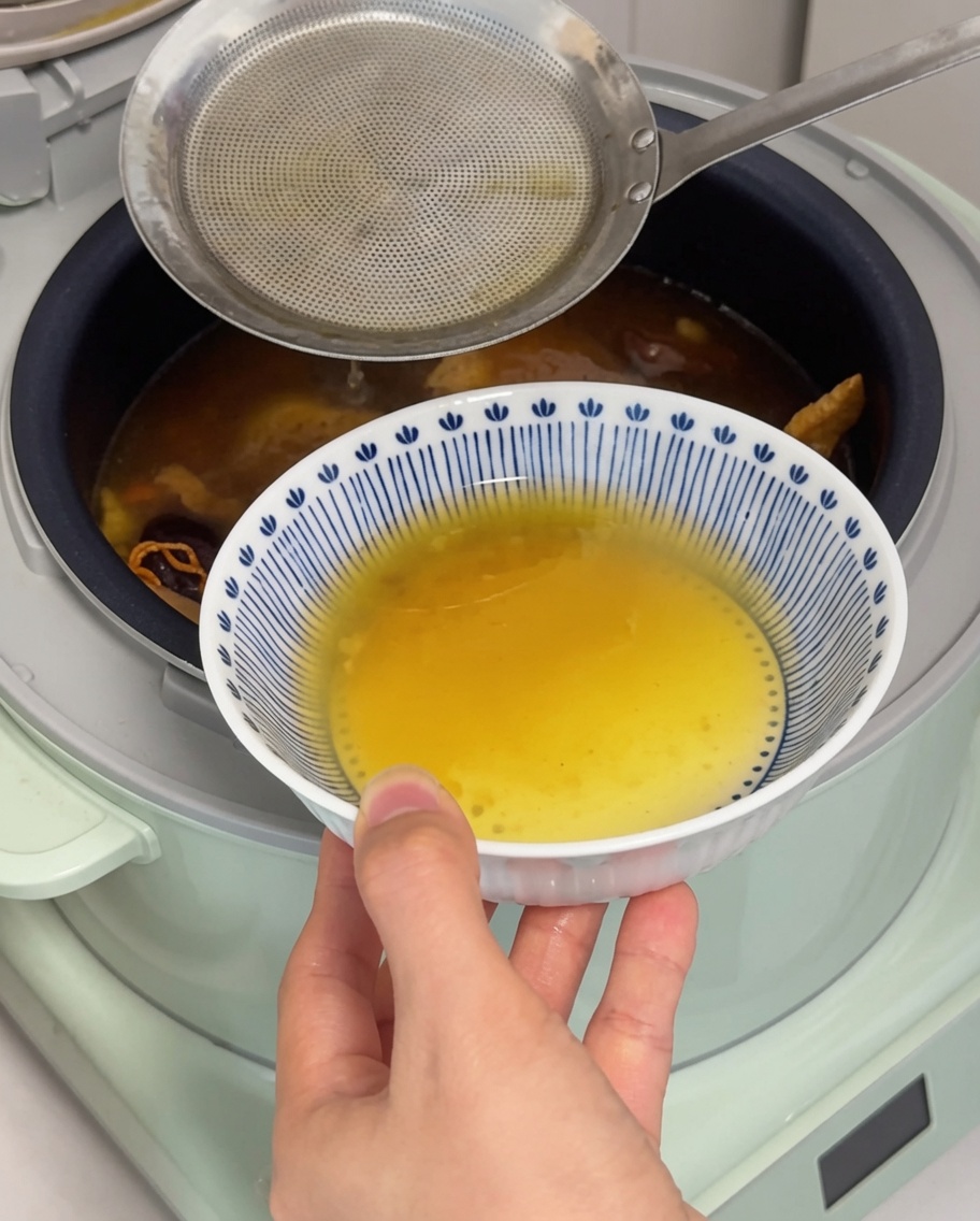 A close-up view of a metal ladle skimming a layer of golden chicken fat from the surface of the soup in a rice cooker.