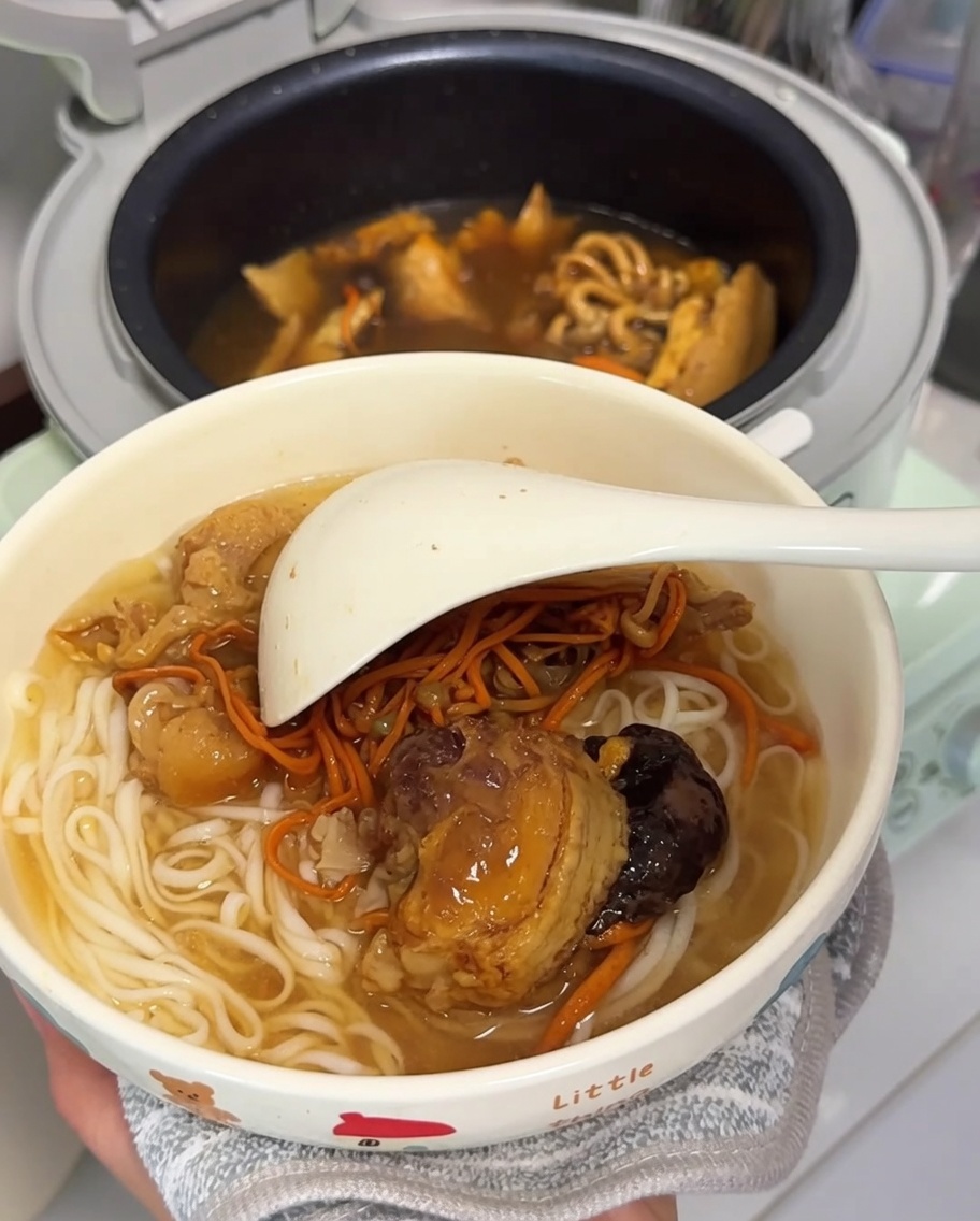 A bowl filled with cooked white noodles and pieces of chicken and dried mushrooms, with a large white ladle resting inside, ready to be served.