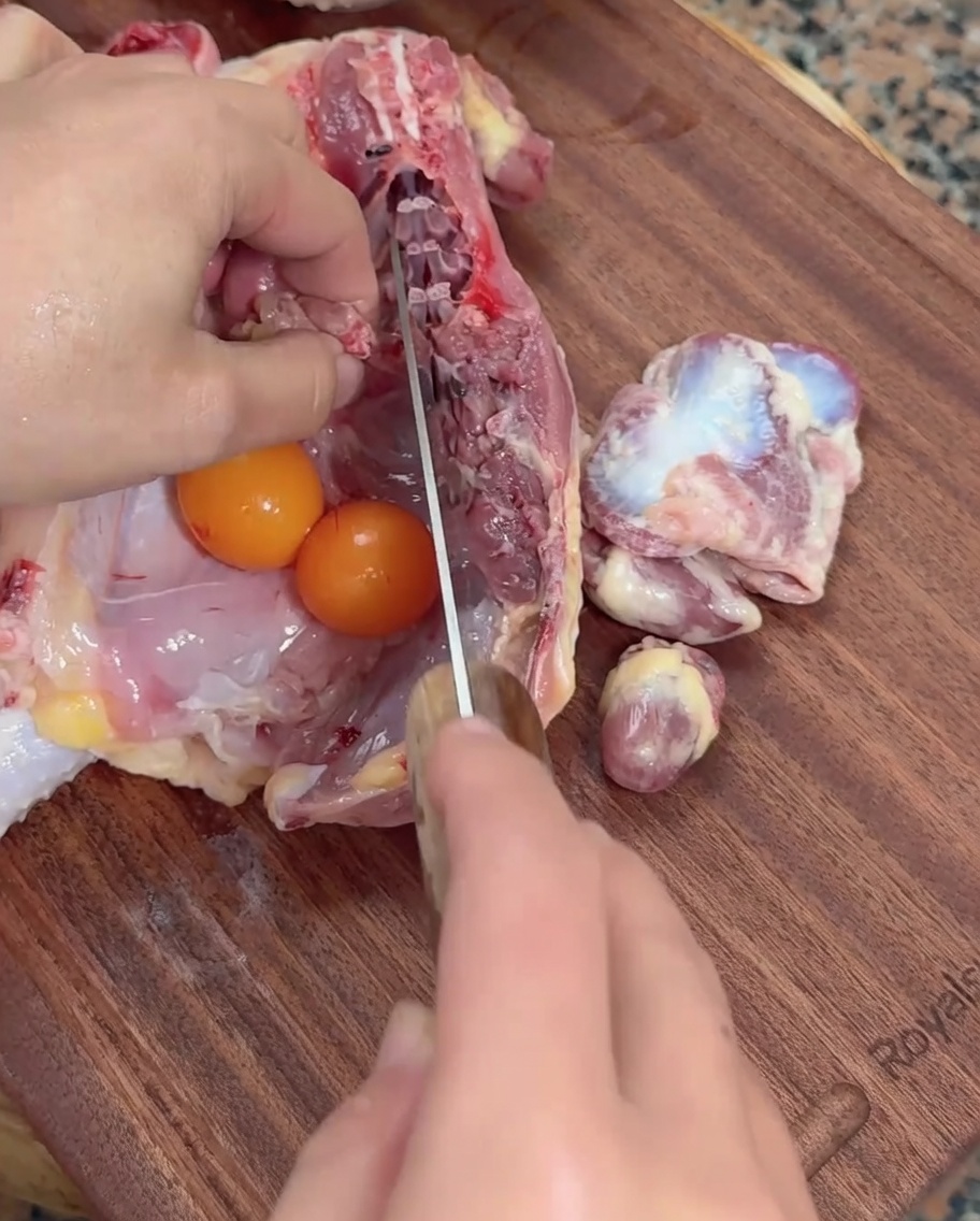 Hands using a cleaver to score raw chicken giblets alongside unlaid chicken eggs on a wooden cutting board.