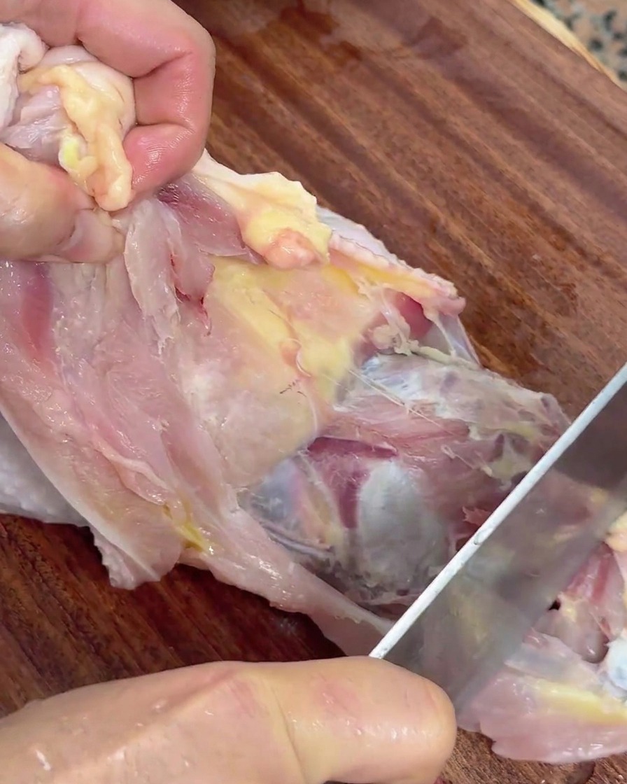 Hands holding a raw chicken and using a knife to slice the meat away from the carcass.