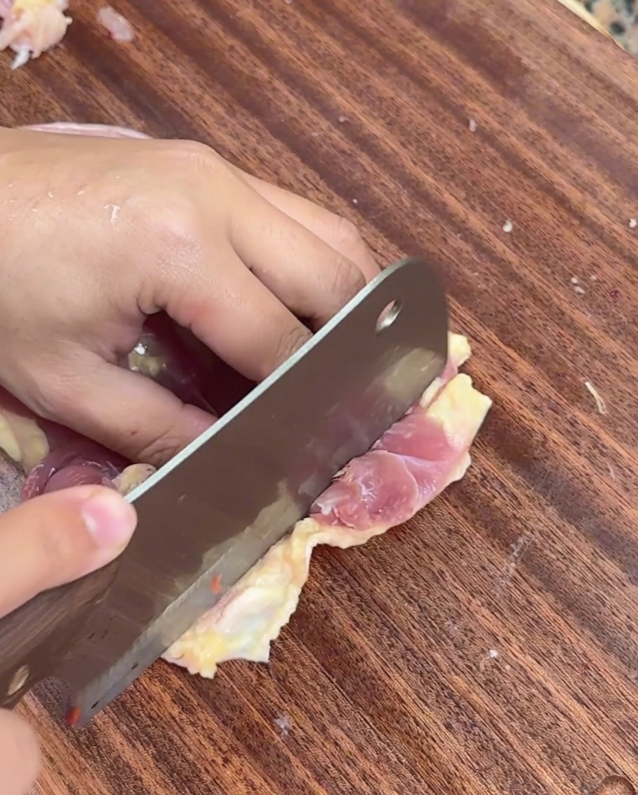 A hand using a cleaver to slice deboned raw chicken meat into thin strips on a wooden board.