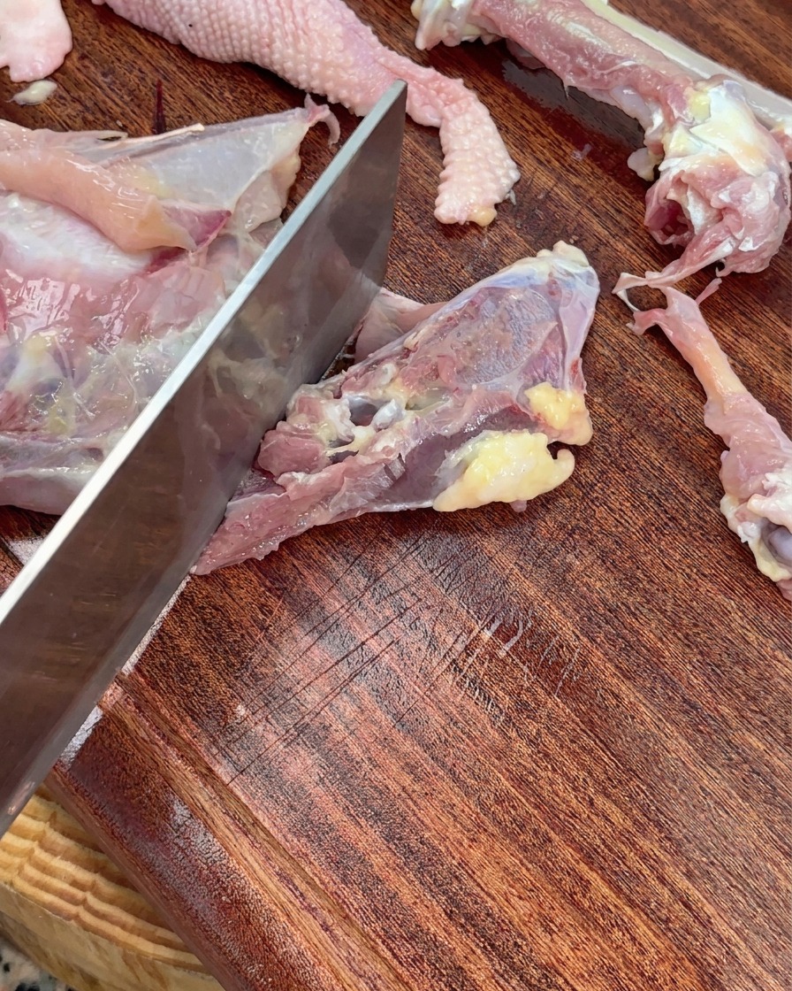 A cleaver chopping a raw chicken skeleton into large pieces on a wooden cutting board.