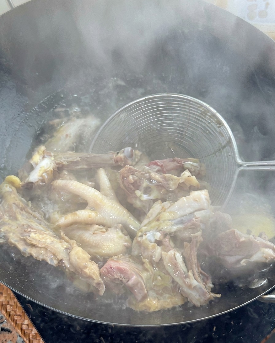 A metal wire strainer lifting blanched chicken bones out of a wok of boiling water.