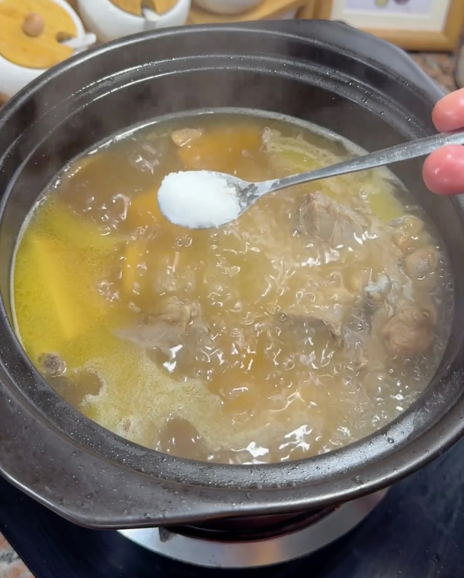 Adding a spoonful of white salt to a simmering golden chicken broth.
