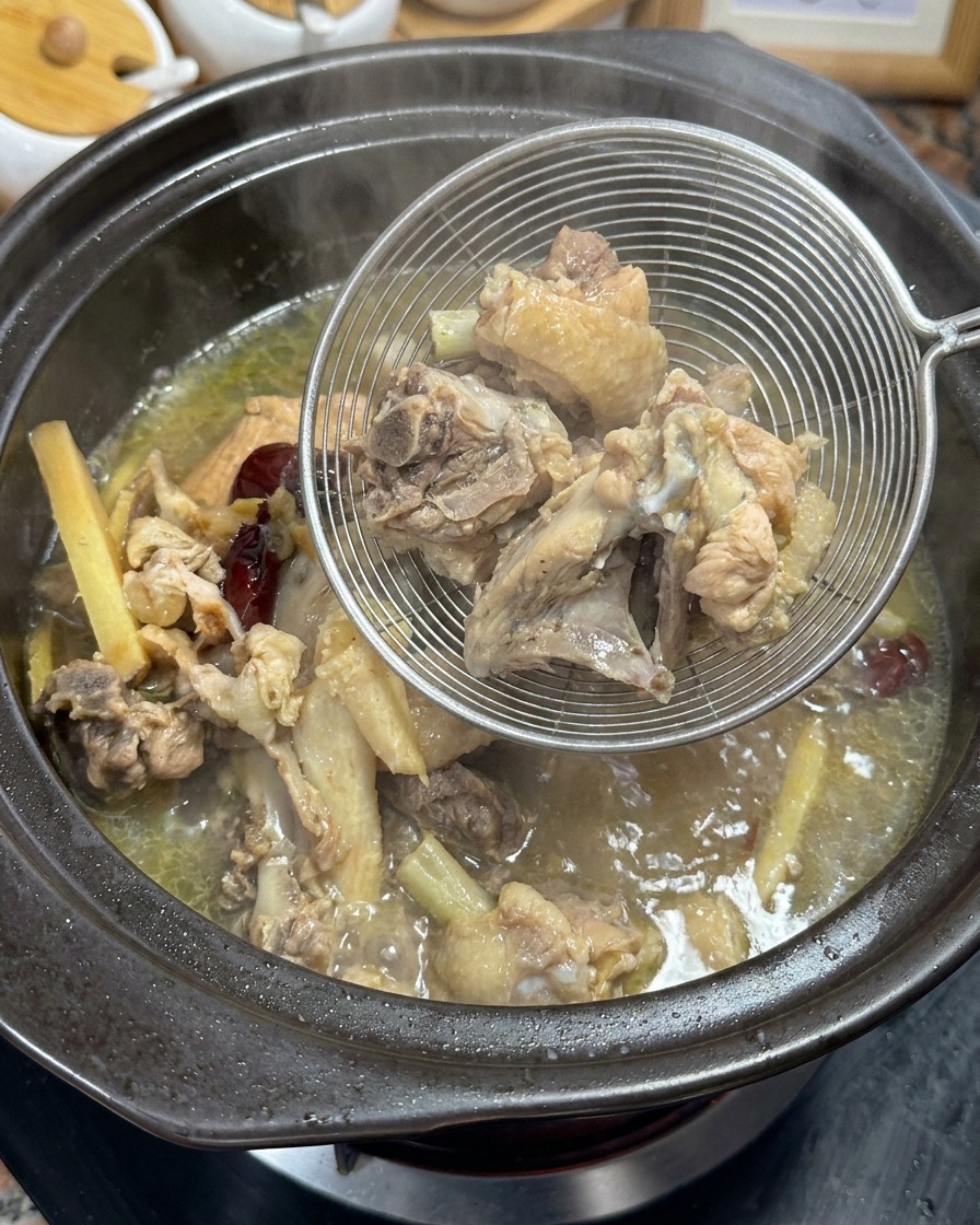 Lifting out boiled chicken bones and aromatics from golden broth using a metal strainer.