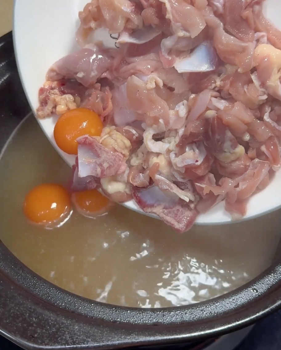 Thinly sliced raw chicken meat and yellow yolks being added into a pot of boiling golden chicken broth.
