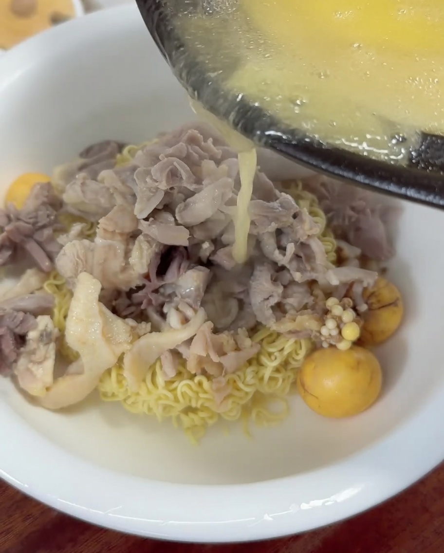 Hot, golden chicken broth being poured from a ladle into a bowl of noodles and chicken.