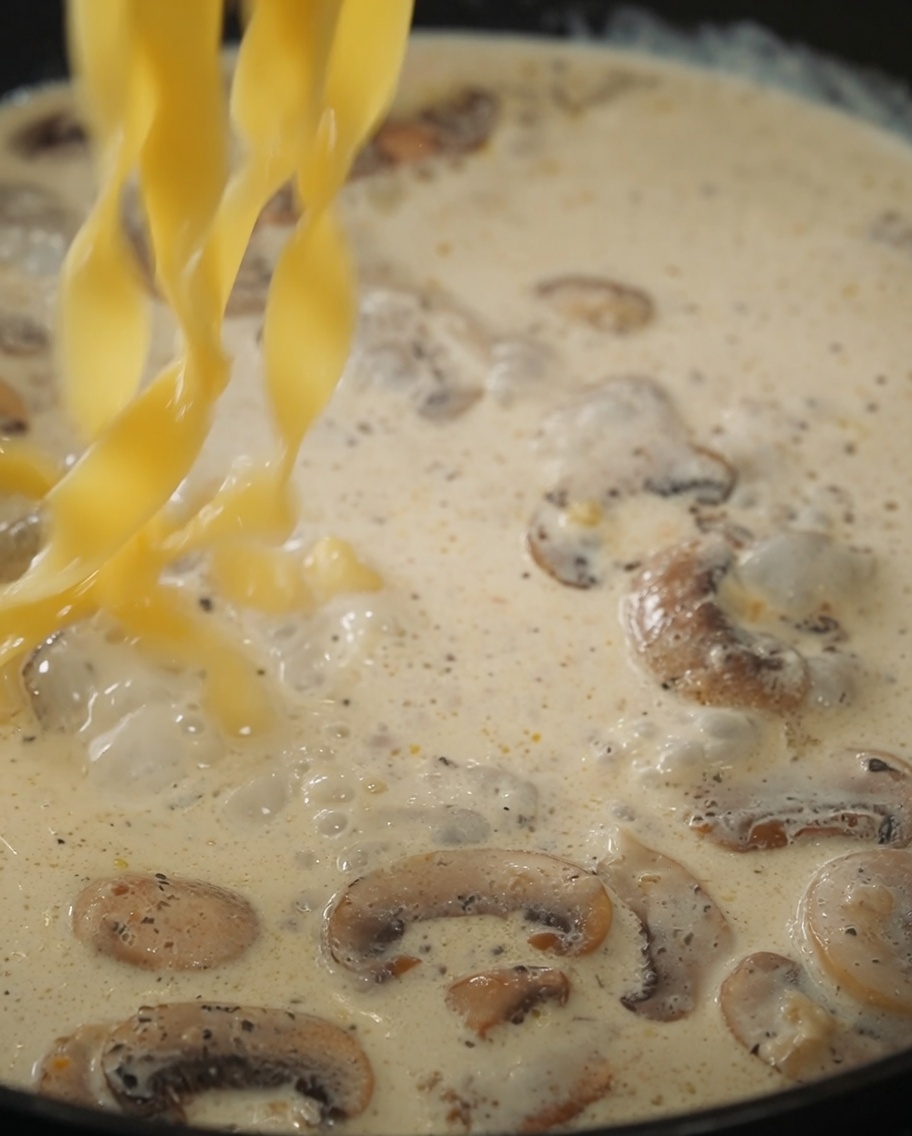 Pasta and black truffle sauce being mixed into the creamy mushroom sauce in a pan.