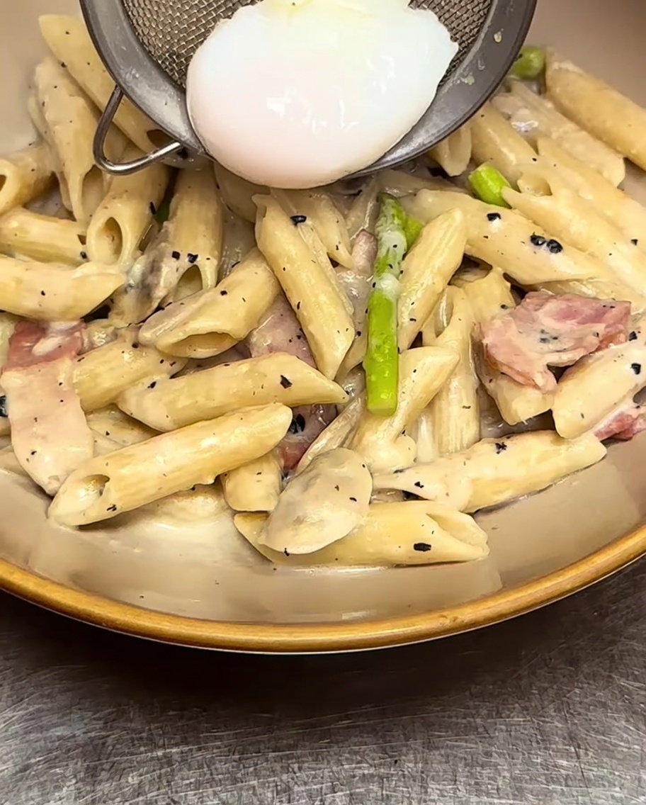 Creamy penne pasta being plated in a bowl with a poached egg placed carefully on top.