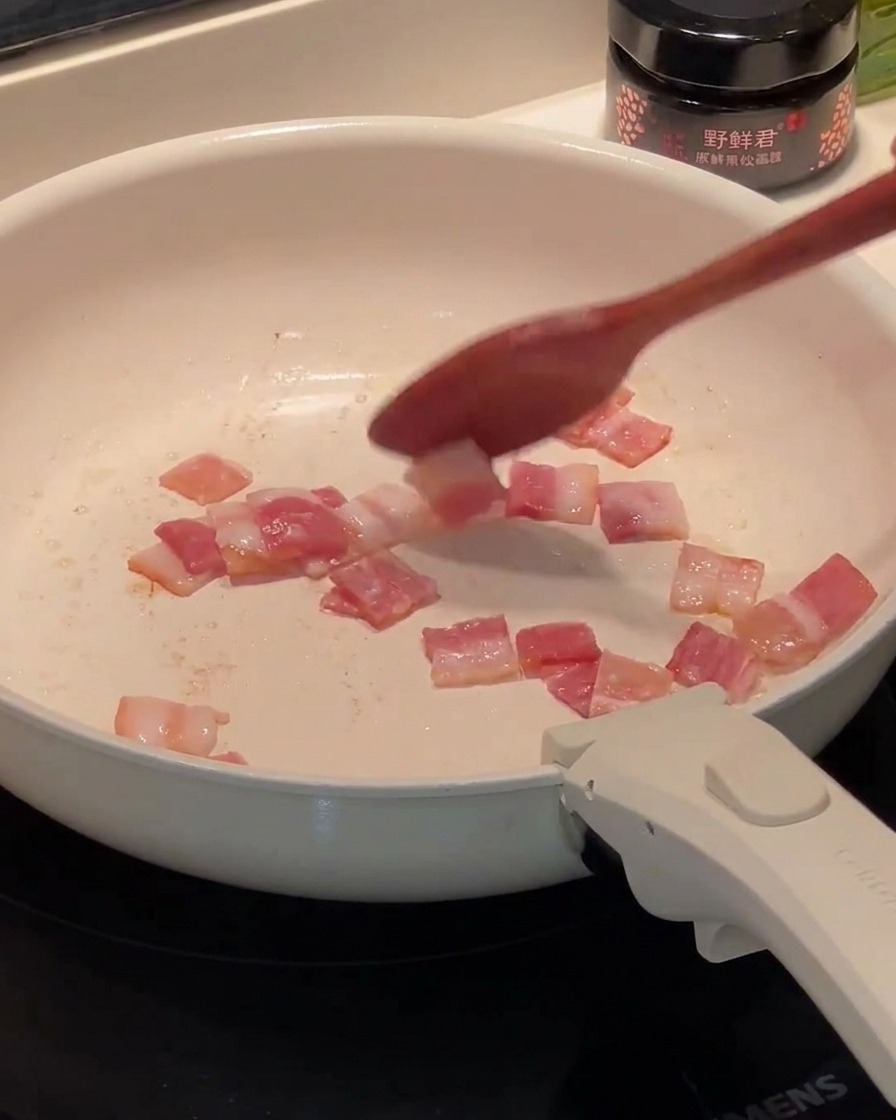 Chopped bacon frying in a white pan, with fat starting to pool at the bottom.