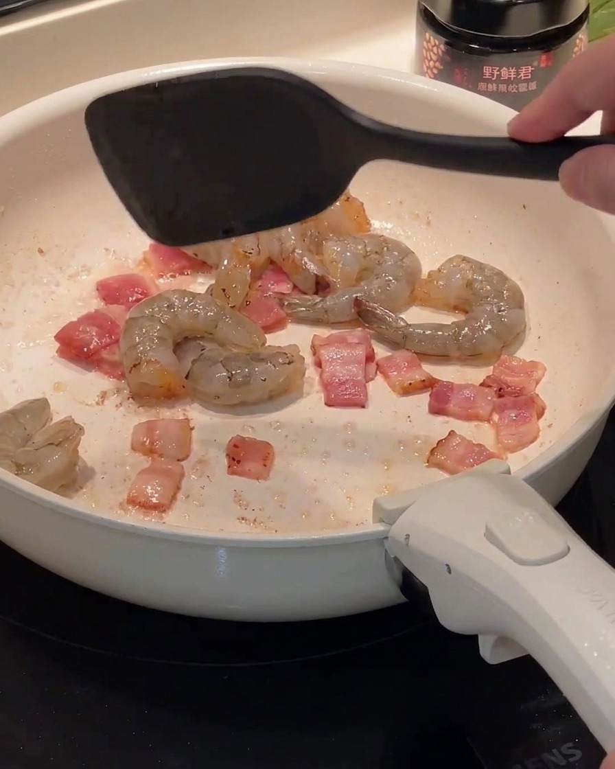 Raw shrimp being added to a pan containing cooked bacon pieces.