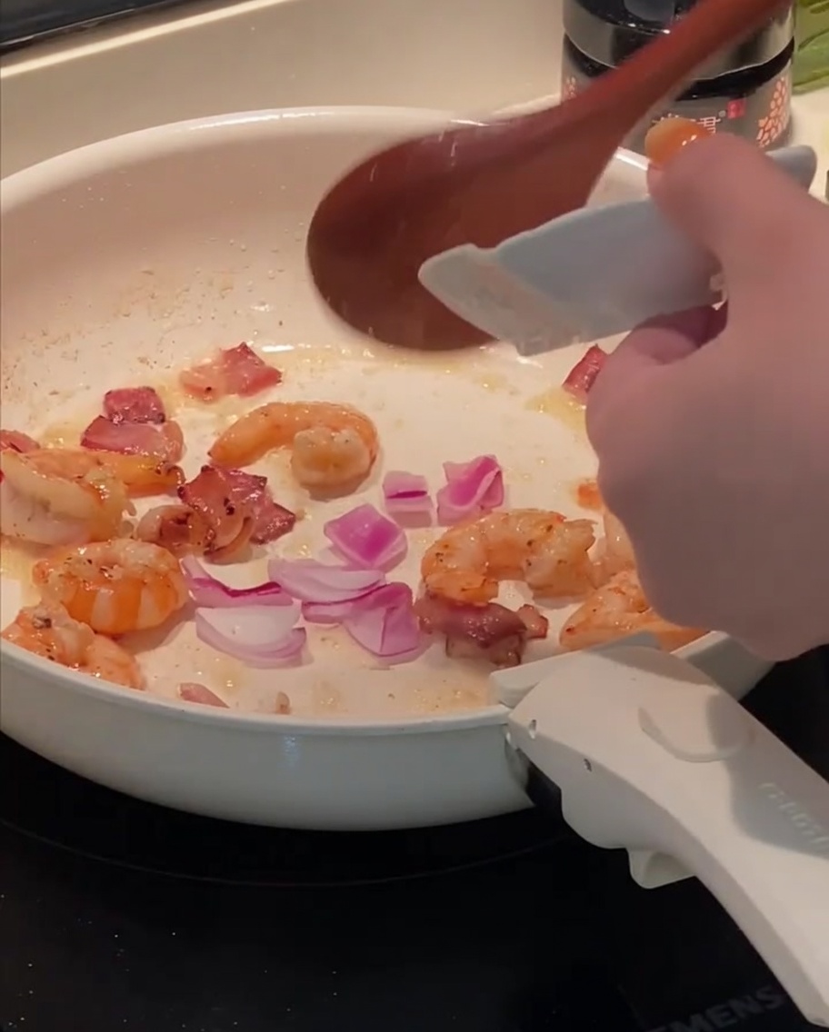Chopped red onion being added to a pan with sautéed bacon and shrimp.