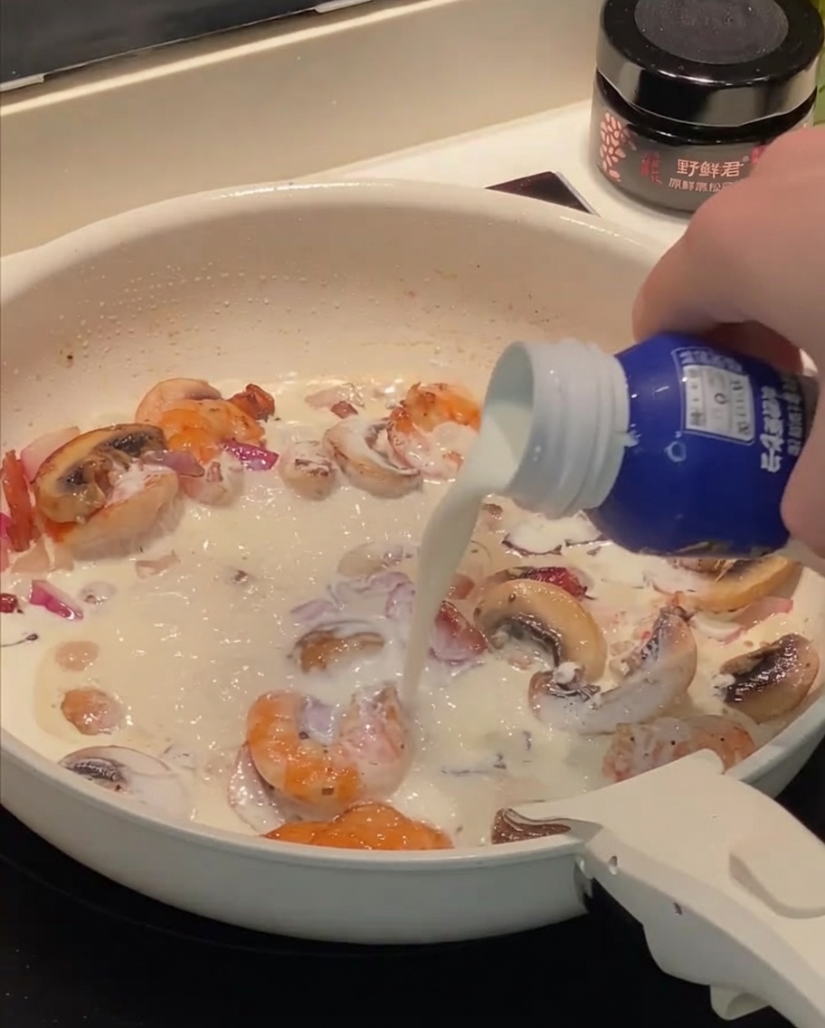 Heavy cream and milk being poured into the pan with sauteed shrimp, bacon, and mushrooms.