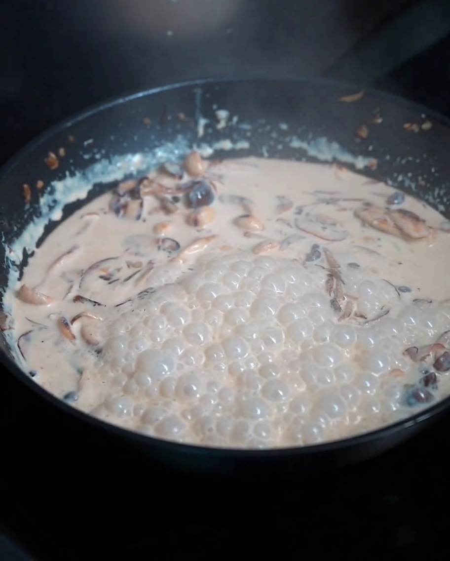 Boiled pasta noodles being tossed into a rich, creamy mushroom sauce in a pan.