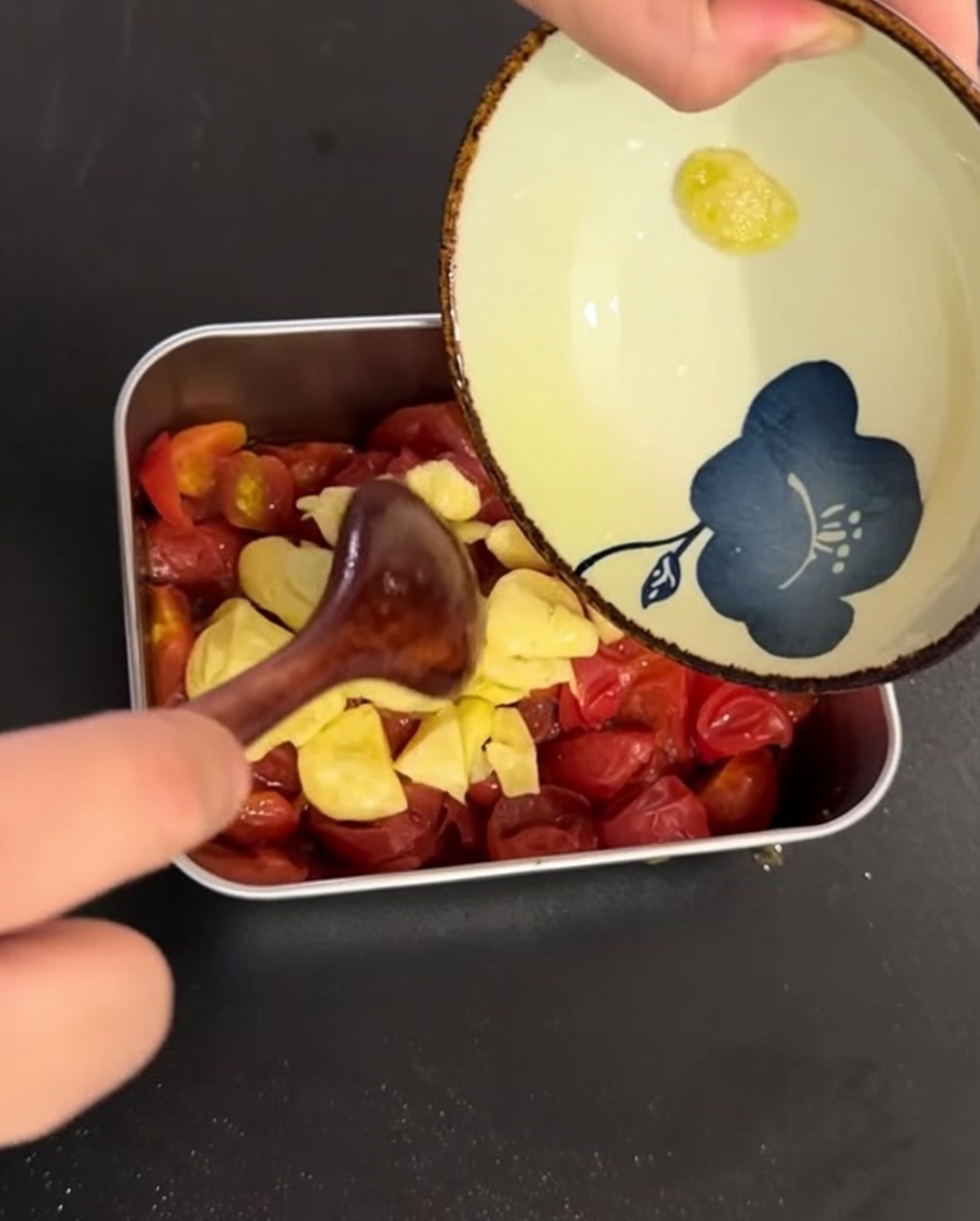 The garlic-infused olive oil and sliced garlic being poured over the cooked cherry tomatoes in a metal dish.