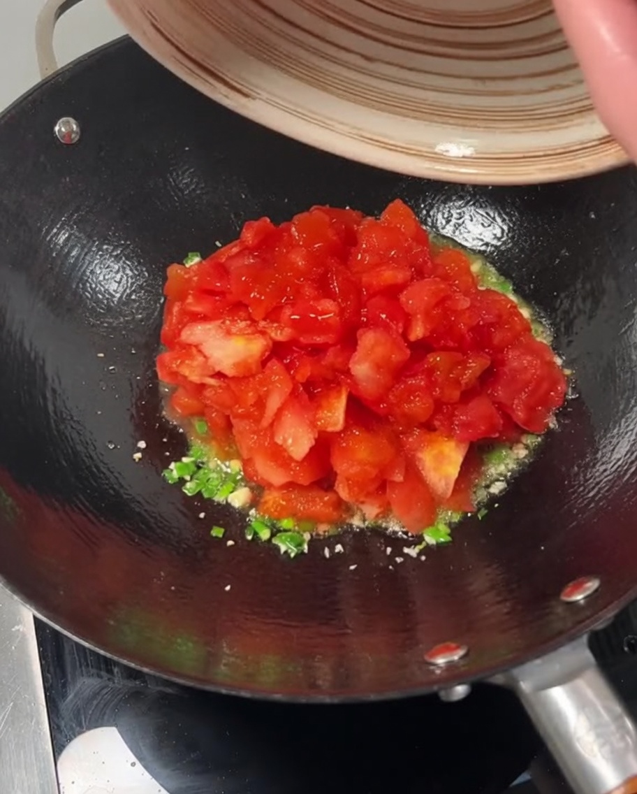 Diced tomatoes being poured from a bowl into a wok over sautéed green onions and garlic.