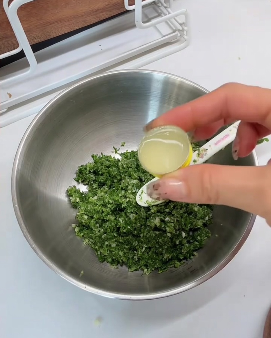 Lemon juice being poured from a small measuring spoon into a metal bowl containing the minced green mixture.
