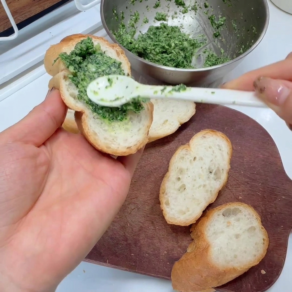 Spreading a bright green garlic and parsley mixture onto slices of baguette with a spoon.