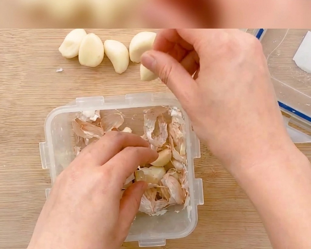 Peeled garlic cloves being sliced into thin pieces on a wooden board.