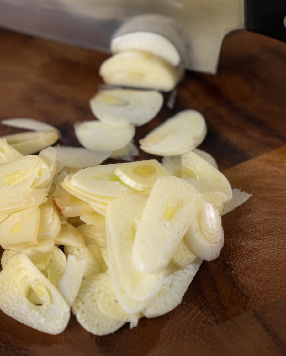Thinly sliced garlic cloves piled on a wooden cutting board next to a chefs knife blade.