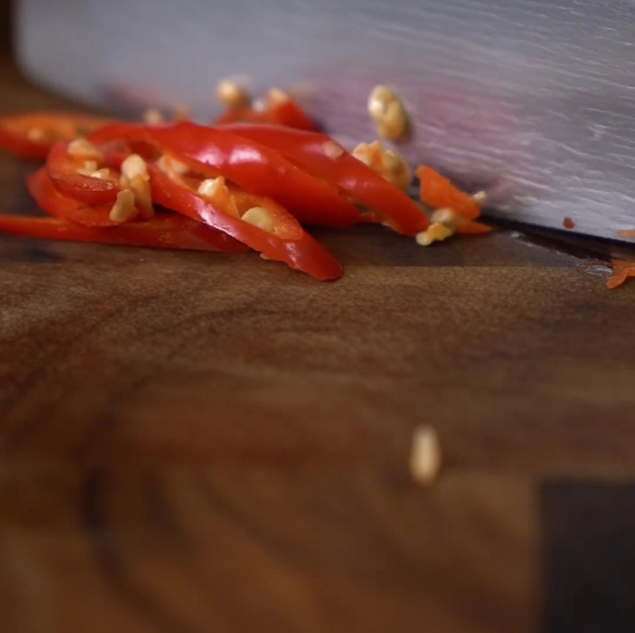 A close-up of a knife slicing bright red chili peppers into small rounds on a dark wooden cutting board.