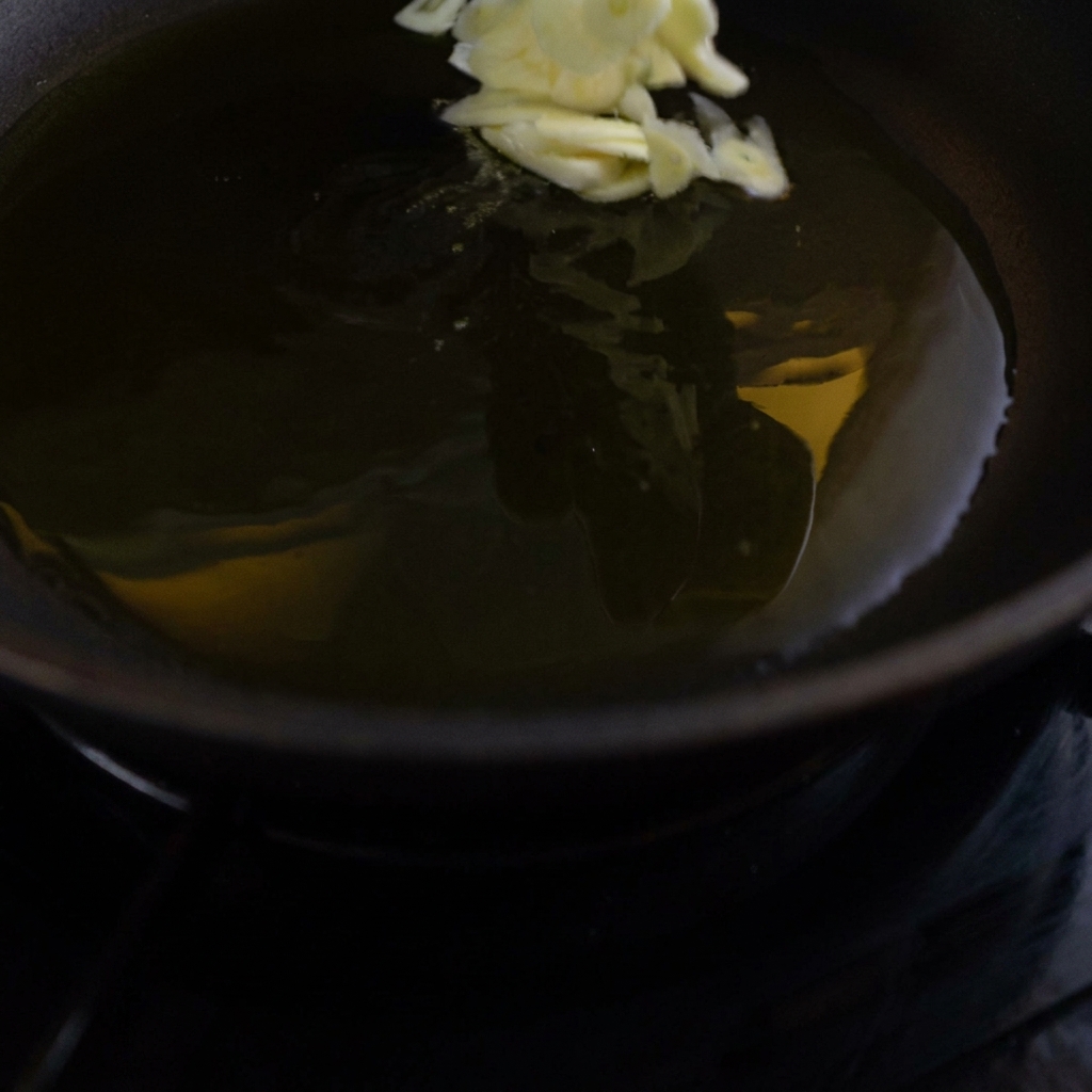 Sliced garlic chips being dropped into a dark frying pan filled with shimmering, hot olive oil.