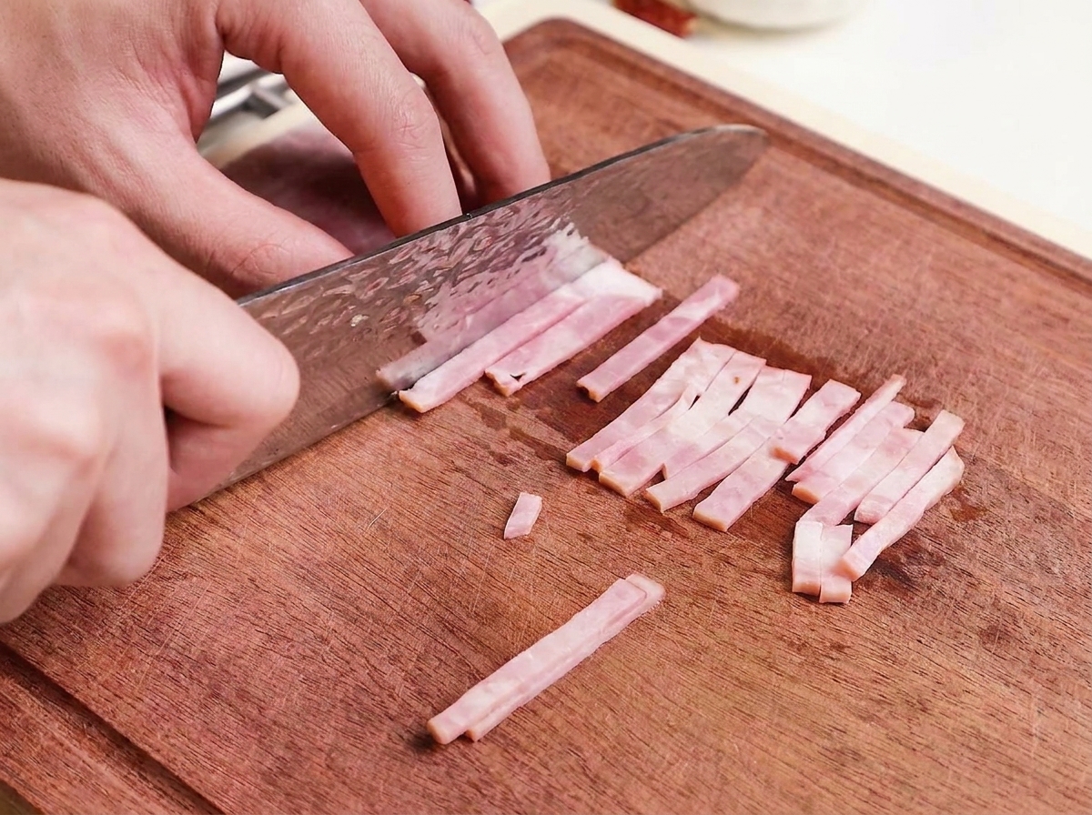 Thin strips of raw bacon being cut on a wooden board.