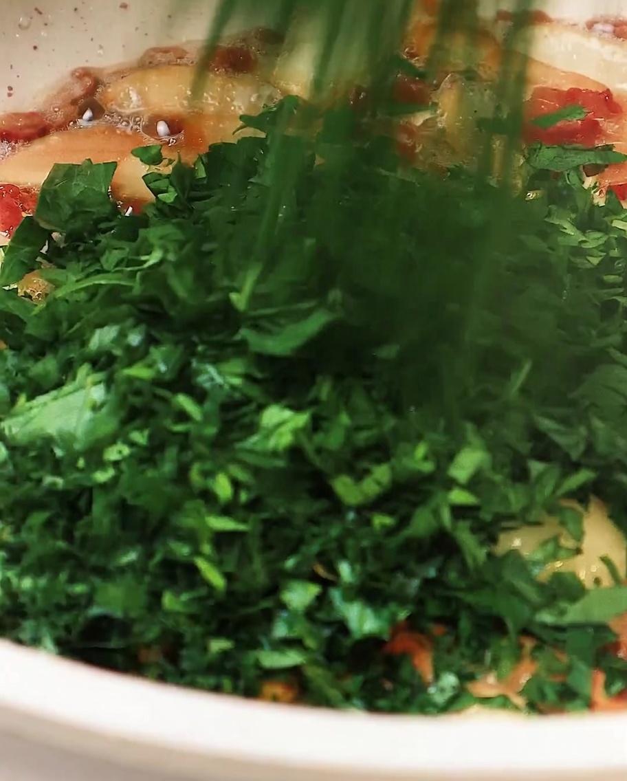 Chopped parsley being added to the pan containing garlic and bacon, lightly stirring to combine.