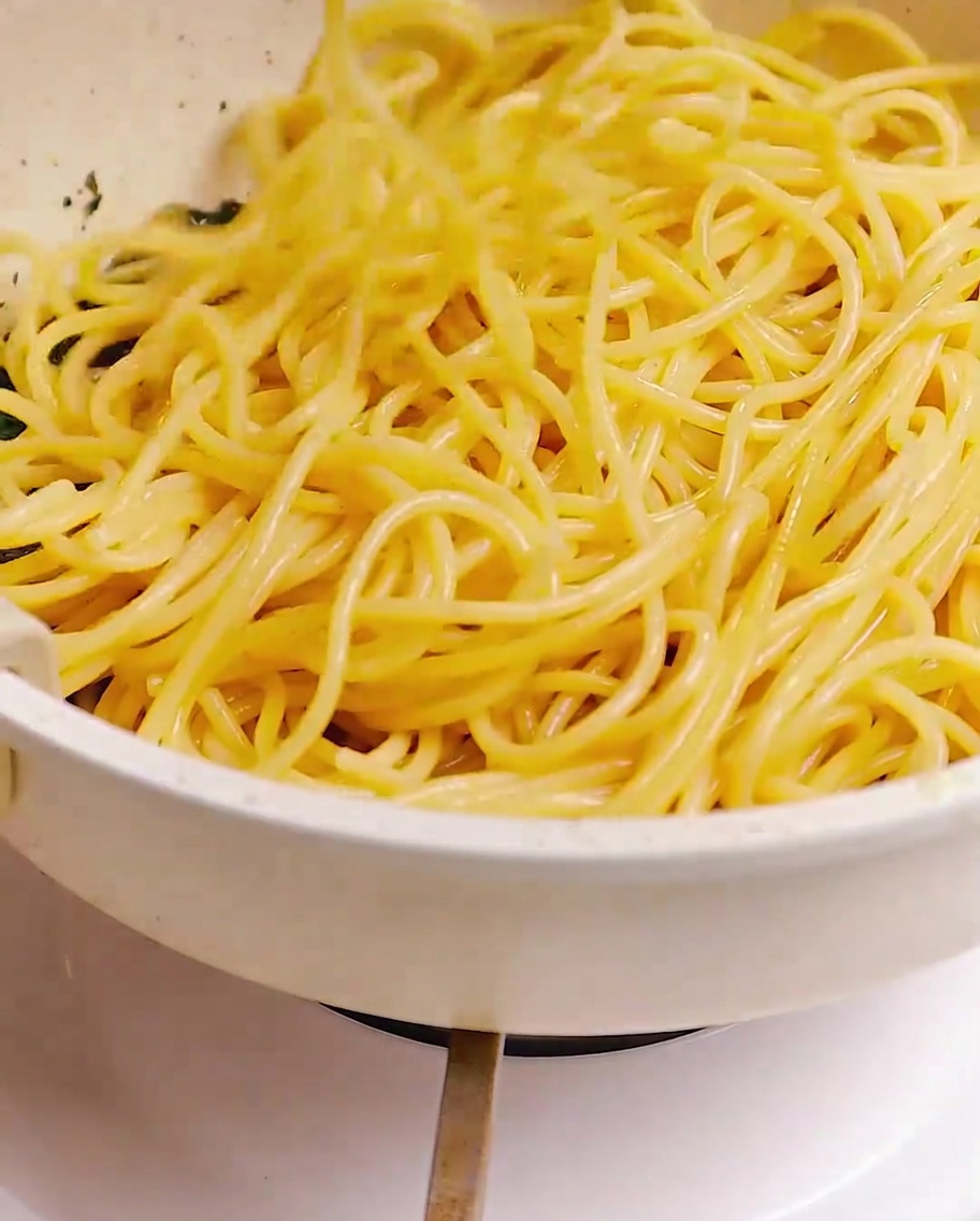 Cooked spaghetti being tossed in a pan with a mixture of bacon, garlic, and herbs using chopsticks.