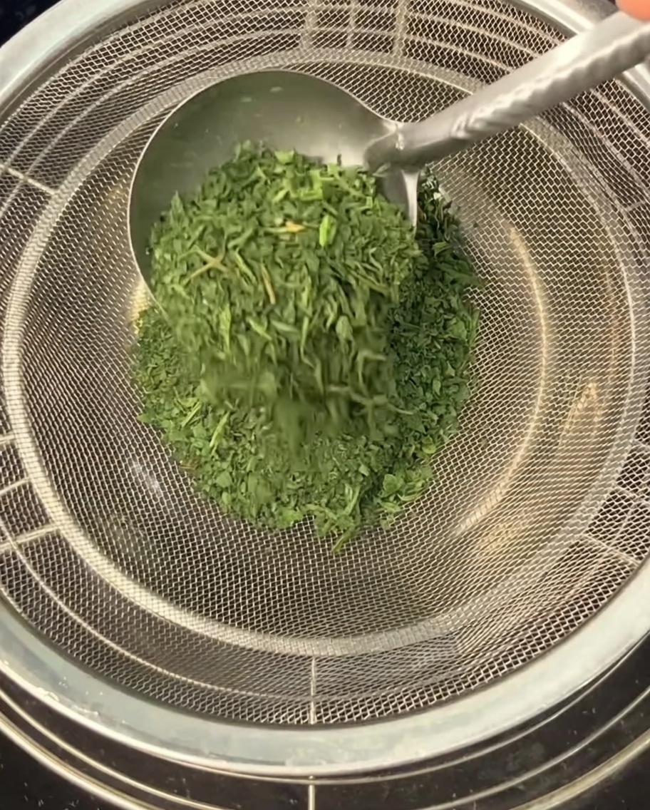 Using a metal spoon to push crushed, dried celery leaves through a fine-mesh strainer into a bowl below.