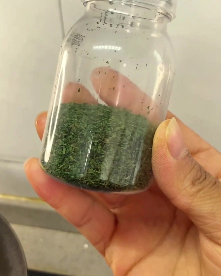 A hand holding a small clear plastic jar partially filled with homemade dried green celery leaf seasoning.